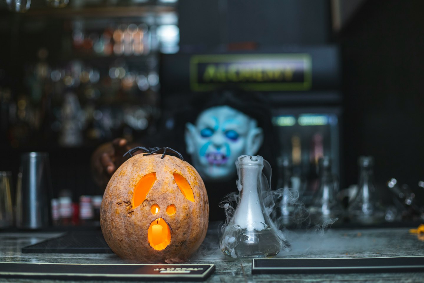 A jack-o'-lantern and a smoky flask sit on a bar.