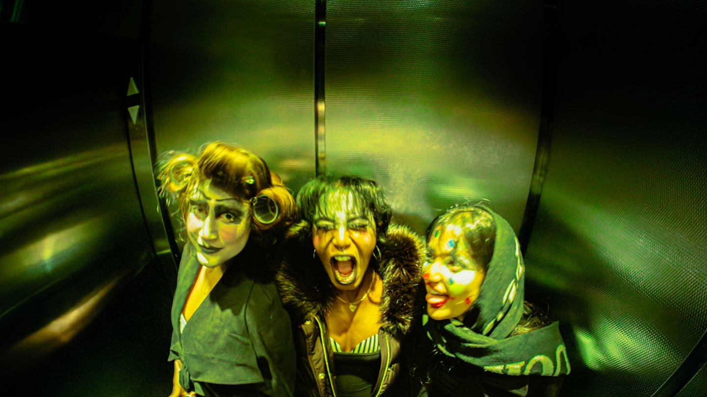 Three performers in theatrical clown makeup pose inside an elevator.
